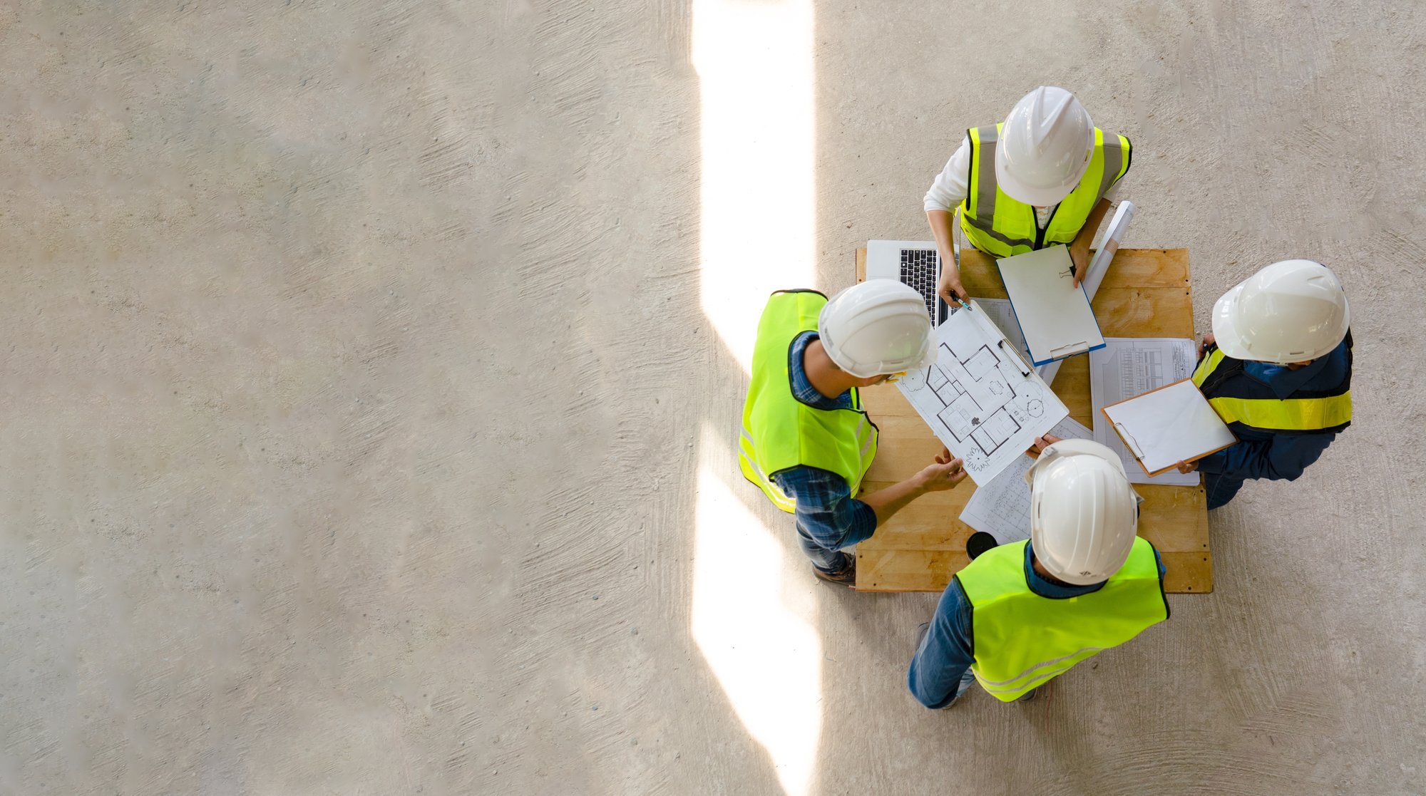 Four construction workers in safety gear reviewing project plans on a table with documents and a laptop.